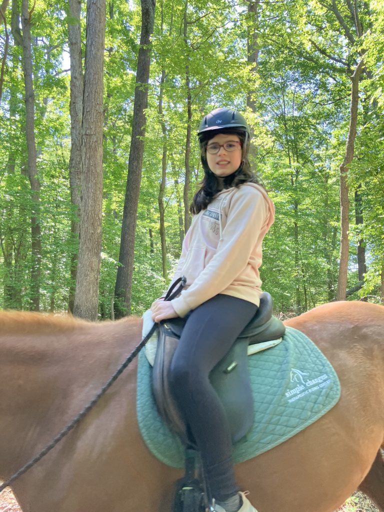 A girl smiles at the camera from where she sits on a chestnut horse. The background is full of bright green leaves on trees.