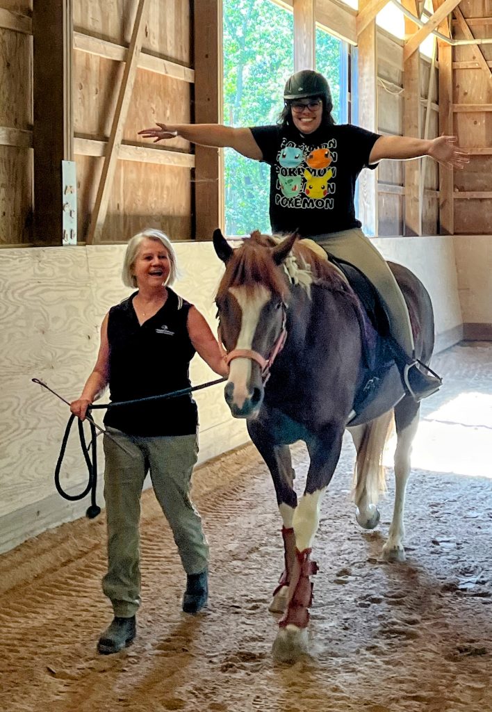 A young woman rides Zelda along the side of the arena. The woman is smiling and holding her arms straight out to the side. Another woman is leading the horse.