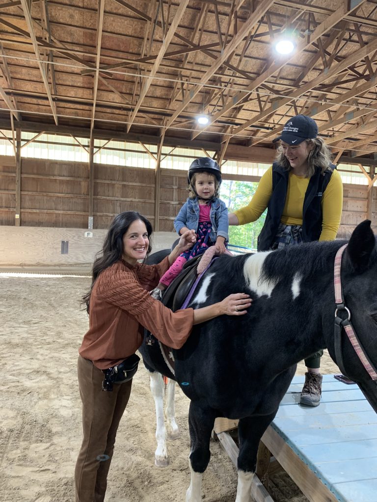 A small child smiles at the camera from where she is sitting on the back of a black and white horse. Adults stand on either side of her. Everyone is smiling and excited.