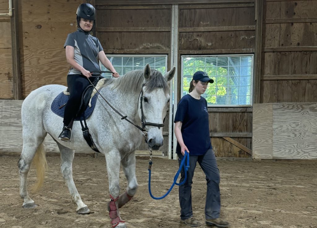 A young woman smiles at the camera as she rides Gemini, a tall grey horse. Another woman is helping to lead Gemini.