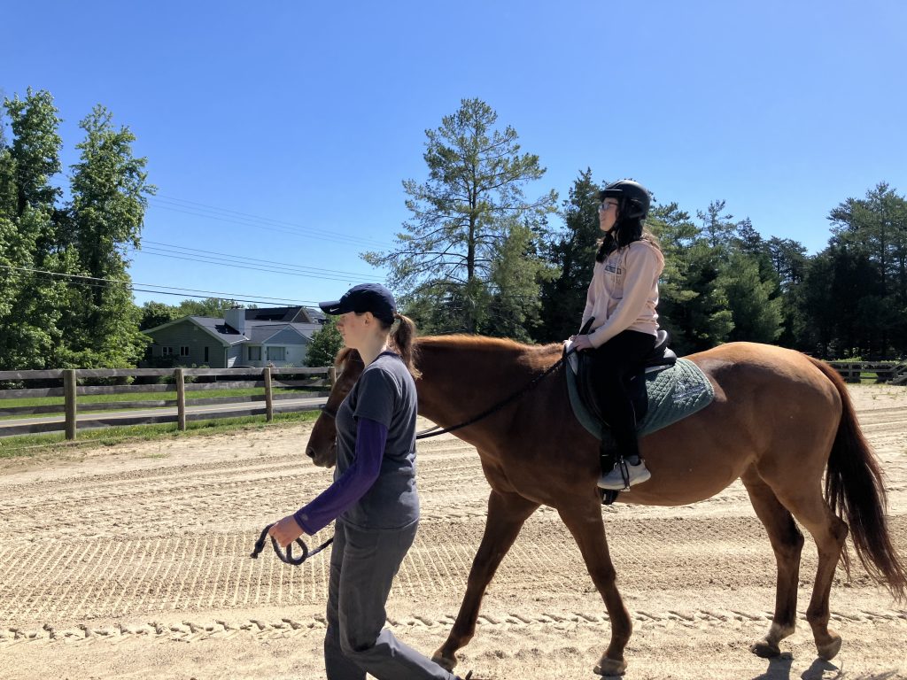 A girl rides Reba in the outdoor arena while a woman leads her.