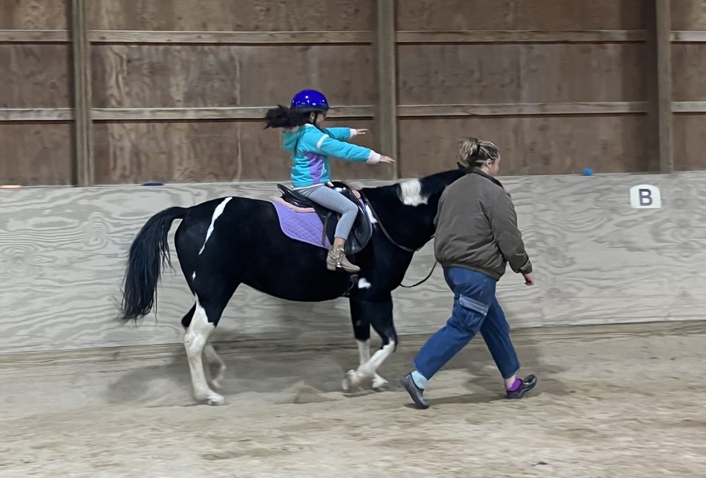 A girl rides Stella at the trot along the arena rail. A leader is guiding Stella so the girl can ride with both arms straight out in front of her.