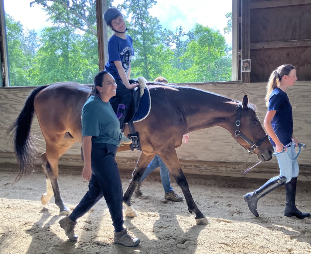A young woman smiles at the camera as she rides Chance alongside the arena wall. Chance is being led by a volunteer and two other volunteers are acting as sidewalkers on either side of the rider.