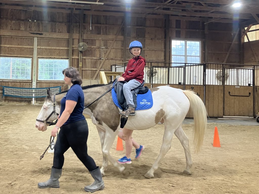 King, a paint horse, is led across the arena by a woman while a child sits on his back and smiles at the camera.