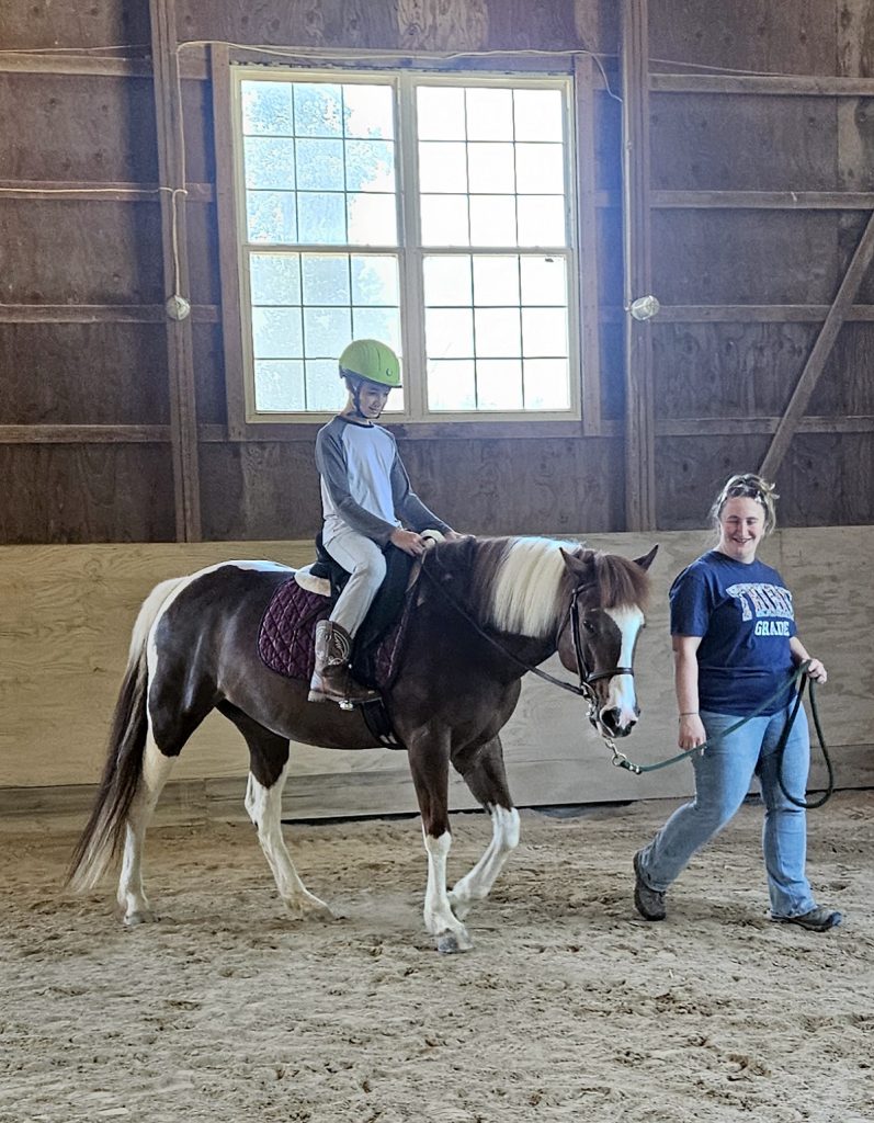 A boy rides Zelda, a brown and white paint mare, in the indoor arena. A woman helps lead Zelda while the boy concentrates on his task.